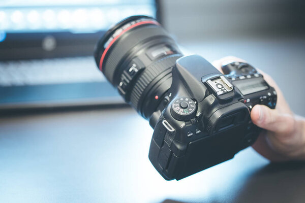 Photographer is holding a professional camera with telephoto lens in his hand, laptop in the blurry background