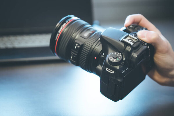 Photographer is holding a professional camera with telephoto lens in his hand, laptop in the blurry background