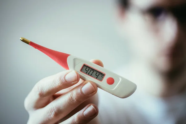 Man holds a red fever thermometer with high temperature in his hand, blurry face