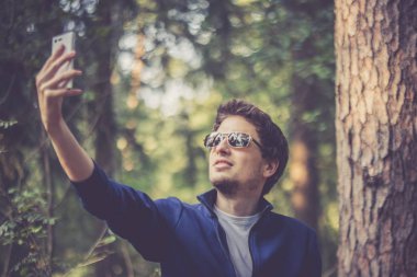 Güneş gözlüklü genç adam parkta selfie çekiyor.