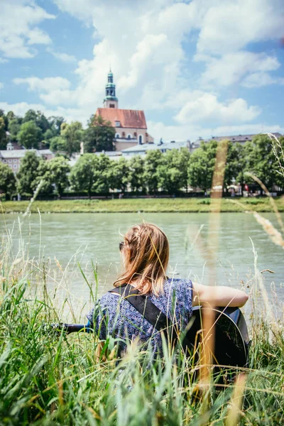 Beautiful young girl is sitting at a river and playing her western guitar