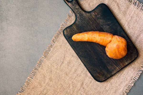 Ugly food. Trendy imperfect carrot on wooden board. Misshapen produce, weird fruits and vegetables, food waste problem concept. Top view, copy space.