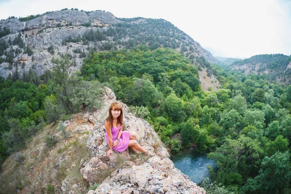 The girl is sitting on a rock. Down the river runs. Behind the rocks and crowns of trees