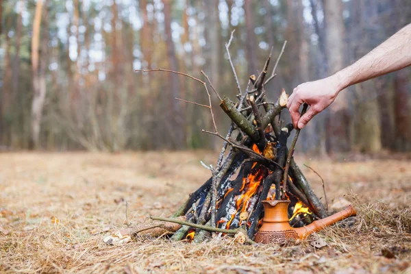 At the stake in the spring forest, a clay Turkish coffee pot is heated against the grass. In the frame one man\'s hand.