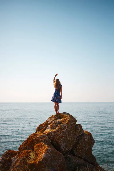 Una chica en un vestido se para en una piedra y mira el mar azul — Foto de Stock