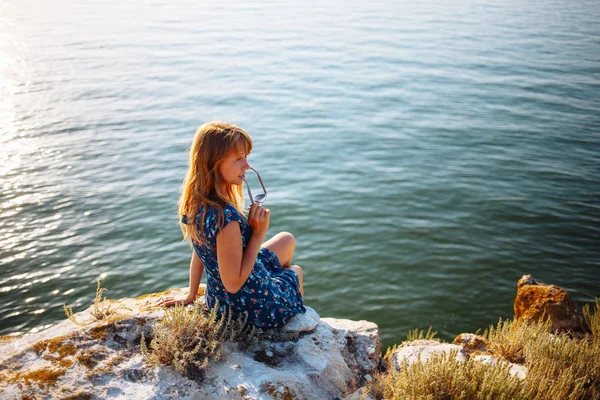 La chica en el vestido azul en la piedra se sienta junto al mar — Foto de Stock