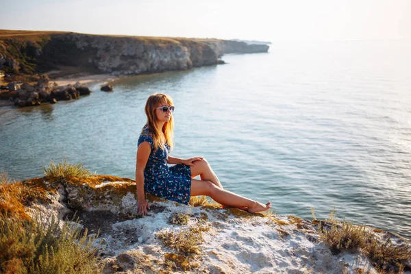 La chica en el vestido azul en la piedra se sienta junto al mar — Foto de Stock