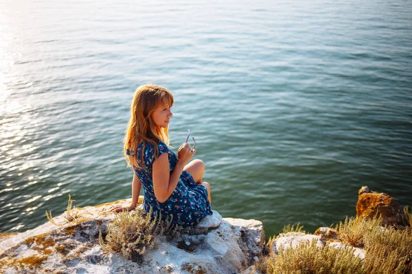 La chica en el vestido azul en la piedra se sienta junto al mar — Foto de Stock