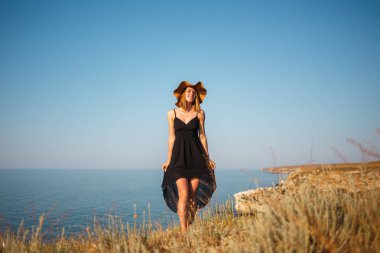 Kayalık bir plajda siyah elbise ve şapkalı kız denize bakıyor.