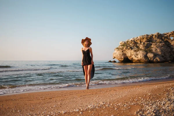 Hermosa chica en un vestido negro y sombrero camina a lo largo de la orilla arenosa del mar al atardecer — Foto de Stock
