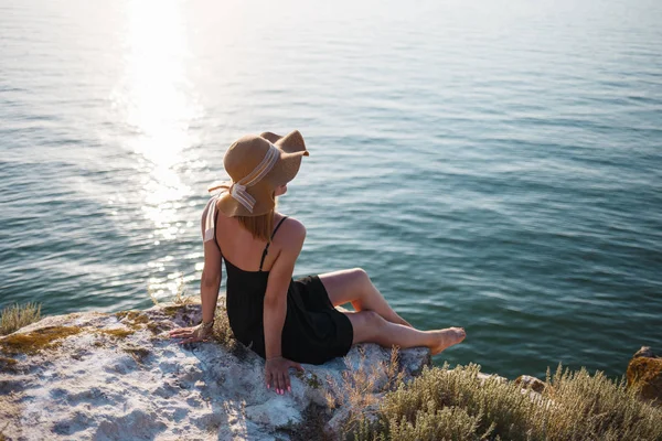 La chica en un vestido negro y sombrero en una playa rocosa mira al mar — Foto de Stock