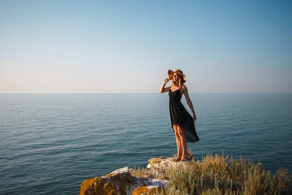 La chica en un vestido negro y sombrero en una playa rocosa mira al mar — Foto de Stock
