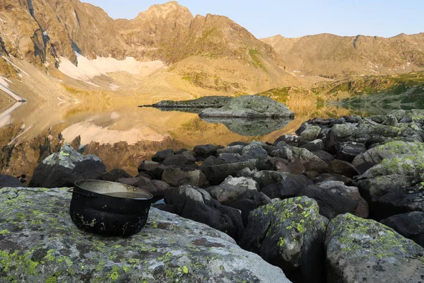 Camping pot with water in the background of mountains mirror reflection in the lake. Hiking motivational image