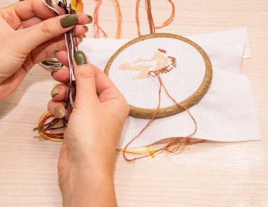 Çemberdeki çapraz dikiş nakışı ahşap bir arka planda yatıyor, kadın elleri çok renkli iplikleri tutuyor.