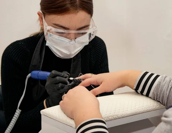 young girl, a manicurist in a mask and glasses, removes the gel polish coating with an apparatus for a client