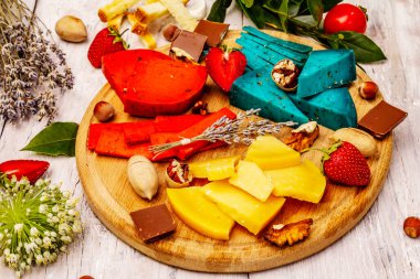 Peynir tabağı. Çeşitli renkte sert Hollanda peynirleri. Mavi ve kırmızı pesto, yıllanmış gouda. Çilek, çikolata, fındık, bitki ve çiçek. Beyaz tahta arka plan, kapat