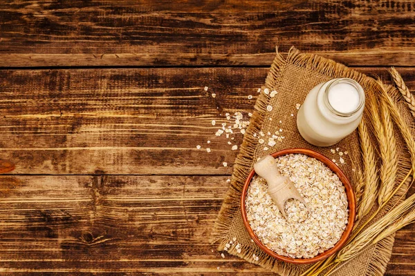Vegan oat milk, non dairy alternative milk. Dry flakes in ceramic bowl, glass bottle, oats. Vintage wooden background, top view