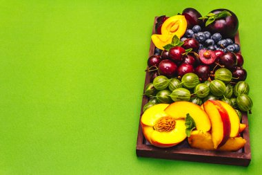 Tabakta çeşitli taze meyveler. Vişne, Bektaşi üzümü, yabanmersini, şeftali dilimleri, erik ve kayısı, kokulu nane. Parlak yeşil arkaplan, kopyalama alanı