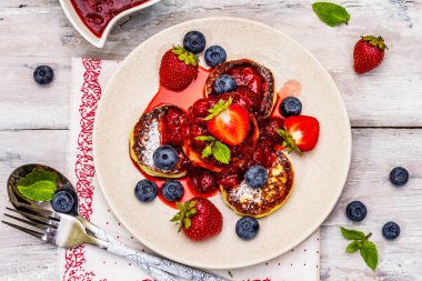 Taze böğürtlen ve çilek soslu süzme peynirli krep. Sağlıklı kahvaltı konsepti. Olgun yaban mersini, kokulu nane yaprakları, çatal bıçak takımı. Tahta kaplama arka plan, üst görünüm