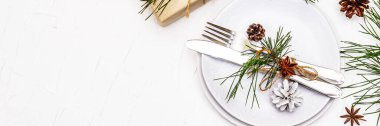 Noel ya da yeni yıl sofrası. Şenlikli çatal bıçak takımı, taze çam dallı seramik tabaklar, koniler, sıfır atık hediye ve yıldız anason. Beyaz macun arkaplanı, pankart biçimi