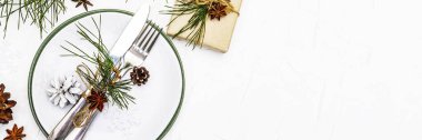 Noel ya da yeni yıl sofrası. Şenlikli çatal bıçak takımı, taze çam dallı seramik tabaklar, koniler, sıfır atık hediye ve yıldız anason. Beyaz macun arkaplanı, pankart biçimi