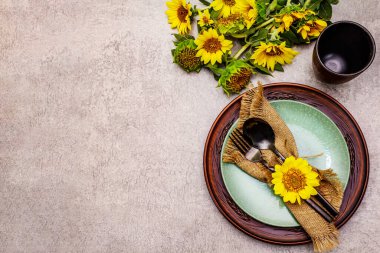 Sonbahar ve Şükran Günü yemeği. Ayçiçeği buketi, seramik tabaklar ve çuval bezi peçete. Taş zeminde siyah çatal bıçak takımı, üst görünüm.