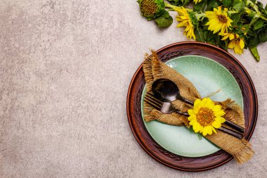 Sonbahar ve Şükran Günü yemeği. Ayçiçeği buketi, seramik tabaklar ve çuval bezi peçete. Taş zeminde siyah çatal bıçak takımı, üst görünüm.