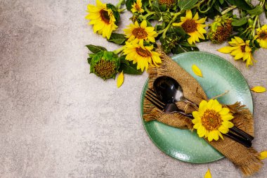Sonbahar ve Şükran Günü yemeği. Ayçiçeği buketi, seramik tabak ve çuval bezi peçete. Taş zeminde siyah çatal bıçak takımı, üst görünüm.