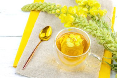 Doğal ahşap zemin üzerinde cam bir bardak Mullein bitkisel çayı ve keten kumaş üzerinde taze sarı Verbascum thapsus çiçekleri. Çevre sağlığı sahnesi, alternatif yaşam tarzı, kopyalama alanı