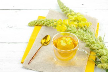 Doğal ahşap zemin üzerinde cam bir bardak Mullein bitkisel çayı ve keten kumaş üzerinde taze sarı Verbascum thapsus çiçekleri. Çevre sağlığı sahnesi, alternatif yaşam tarzı, kopyalama alanı