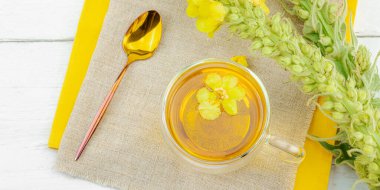 Doğal ahşap zemin üzerinde cam bir bardak Mullein bitkisel çayı ve keten kumaş üzerinde taze sarı Verbascum thapsus çiçekleri. Çevre sağlığı sahnesi, alternatif yaşam tarzı, afiş biçimi