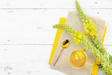 Doğal ahşap zemin üzerinde cam bir bardak Mullein bitkisel çayı ve keten kumaş üzerinde taze sarı Verbascum thapsus çiçekleri. Çevre sağlığı sahnesi, alternatif yaşam tarzı, üst görünüm