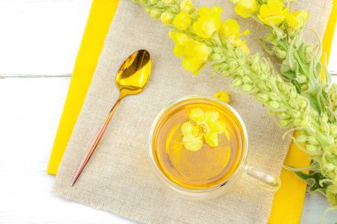 Doğal ahşap zemin üzerinde cam bir bardak Mullein bitkisel çayı ve keten kumaş üzerinde taze sarı Verbascum thapsus çiçekleri. Çevre sağlığı sahnesi, alternatif yaşam tarzı, üst görünüm