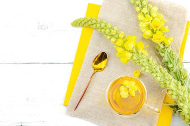 Doğal ahşap zemin üzerinde cam bir bardak Mullein bitkisel çayı ve keten kumaş üzerinde taze sarı Verbascum thapsus çiçekleri. Çevre sağlığı sahnesi, alternatif yaşam tarzı, üst görünüm