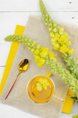 Doğal ahşap zemin üzerinde cam bir bardak Mullein bitkisel çayı ve keten kumaş üzerinde taze sarı Verbascum thapsus çiçekleri. Çevre sağlığı sahnesi, alternatif yaşam tarzı, üst görünüm