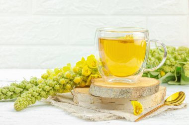 Doğal ahşap zemin üzerinde cam bir bardak Mullein bitkisel çayı ve keten kumaş üzerinde taze sarı Verbascum thapsus çiçekleri. Çevre sağlığı sahnesi, alternatif yaşam tarzı, kopyalama alanı