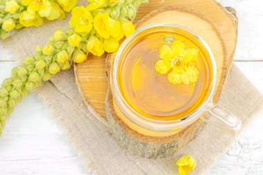 Doğal ahşap zemin üzerinde cam bir bardak Mullein bitkisel çayı ve keten kumaş üzerinde taze sarı Verbascum thapsus çiçekleri. Çevre sağlığı sahnesi, alternatif yaşam tarzı, üst görünüm