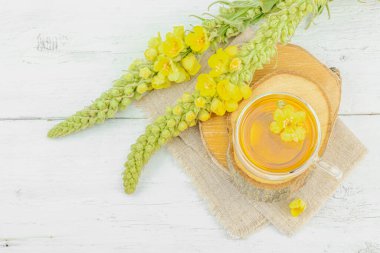 Doğal ahşap zemin üzerinde cam bir bardak Mullein bitkisel çayı ve keten kumaş üzerinde taze sarı Verbascum thapsus çiçekleri. Çevre sağlığı sahnesi, alternatif yaşam tarzı, üst görünüm