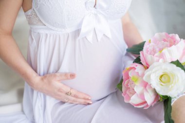 Yakın çekim görüntüsü güzel beyaz elbiseli karnı elleri ile dokunarak ve bir buket peonies tutan hamile kadın. Çiçekli hamile bir kız güzel görüntüsü. Bir çocuk bekliyor. Annelik.