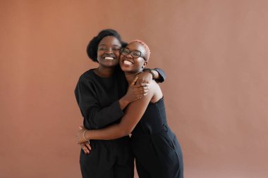Happy african american friends or sisters hugging laughing looking at camera on brown background clipart