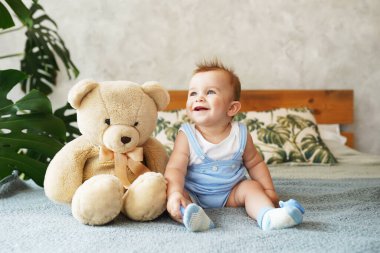 Sevimli küçük çocuk büyük bir yatakta oturuyor, sevimli bir oyuncak ayı ve gülümsüyor.