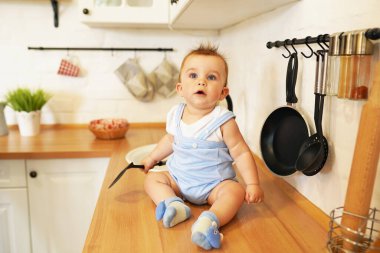 Komik bir erkek bebek mutfak masasında kaşıkla oynar. Küçük bir çocuk metal bir tavanın yanındaki masada oturuyor.