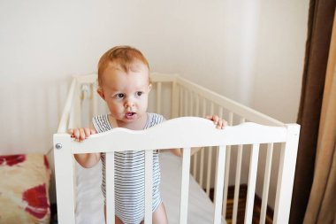 Beyaz yuvarlak bir yatakta duran sevimli bir bebek. Çocuklar için beyaz çocuk odası. Küçük kız beşiğinde durmayı öğreniyor..