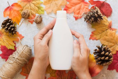 Sonbahar kozmetik ürünleri, şampuanlı beyaz şişe ya da duş jeli kadınların elinde sarı ve kırmızı yapraklı gri arka planda