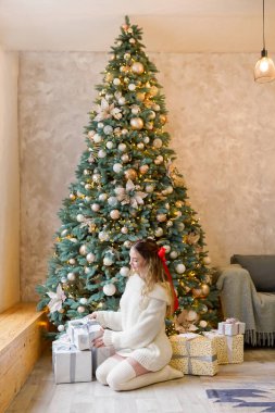 Noel ağacının yanında beyaz kazaklı genç bir kadın. Yeni yılı kutlayan lüks bir kız.