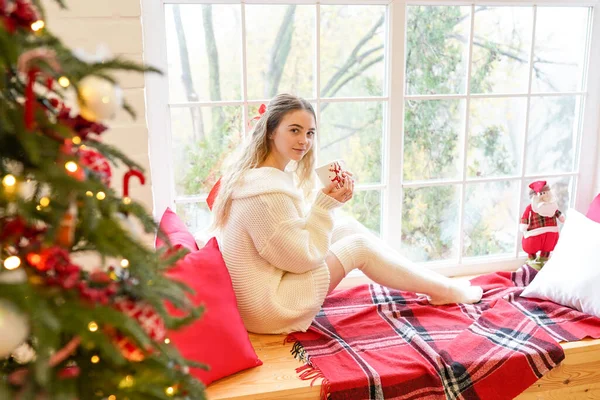 Young woman sitting at home near window with cup of hot coffee wearing knitted warm sweater. Christmas tree with decorations and lights in the room — Stock Photo, Image