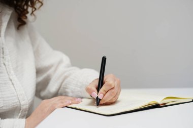 Beyaz gömlekli bir kadın, beyaz bir masanın üzerinde siyah kalemle deftere yazı yazıyor.