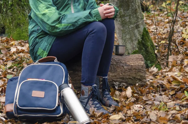 Young woman in a green jacket and black jeans drinking tea from a cup or mug in the autumn forestt. Walk in the woods. Stop for a rest and a snack. Hiking in the fall.