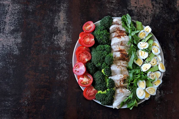 Plate with a keto diet food. A set of products for the ketogenic diet on a plate. Cherry tomatoes, boiled broccoli, steamed chicken breast, salad with arugula, avocado and quail eggs. Keto lunch.