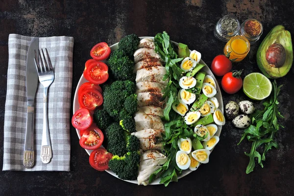 Plate with a keto diet food. A set of products for the ketogenic diet on a plate. Cherry tomatoes, boiled broccoli, steamed chicken breast, salad with arugula, avocado and quail eggs. Keto lunch.